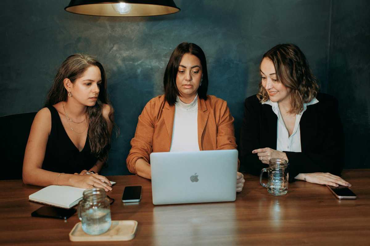 Three women engaged in a work discussion around a laptop in a professional setting.