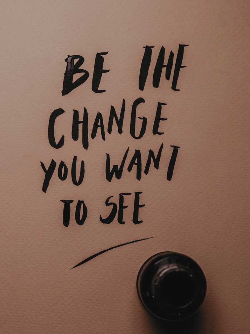 Inspirational message 'Be the change you want to see' in black ink on pink surface.